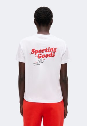 Persoon die van zich af staat, draagt een wit T-shirt met rode tekst "Sporting Goods" en rode short tegen een effen lichte achtergrond.
