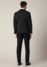Mężczyzna stojący tyłem, ubrany w czarną marynarkę, czarne spodnie, białe mankiety koszuli i czarne buty eleganckie, na neutralnym tle.
