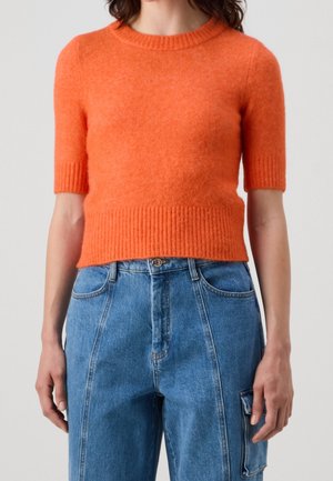 Personne portant un pull à manches courtes orange vif assorti à un jean en denim bleu taille haute avec une poche latérale.
