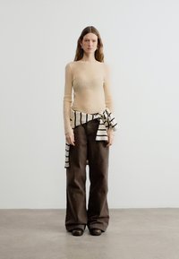 Femme portant un pull côtelé beige clair, un pantalon large marron foncé, des chaussures noires, et un pull rayé blanc et noir noué autour de la taille.