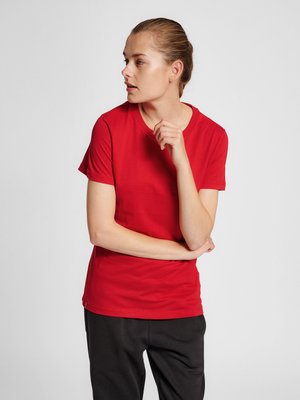 Vrouw met naar achteren getrokken haar, die een rood T-shirt met korte mouwen en zwarte broek draagt, kijkt naar links met gekruiste armen en hand bij haar kin.