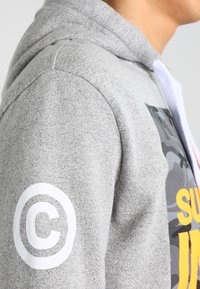 Persona que lleva una sudadera gris con un símbolo de copyright en blanco en la manga y texto amarillo parcialmente visible en el pecho.