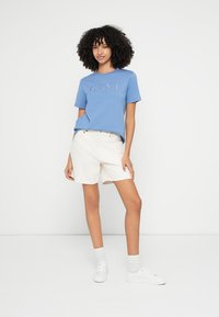 Zils īsroku t-krekls ar reljefa logotipu, kopā ar bēšiem šortiem. Modelis valkā baltas sporta kurpes un zeķes, pozējot pret neitrālu fonu.