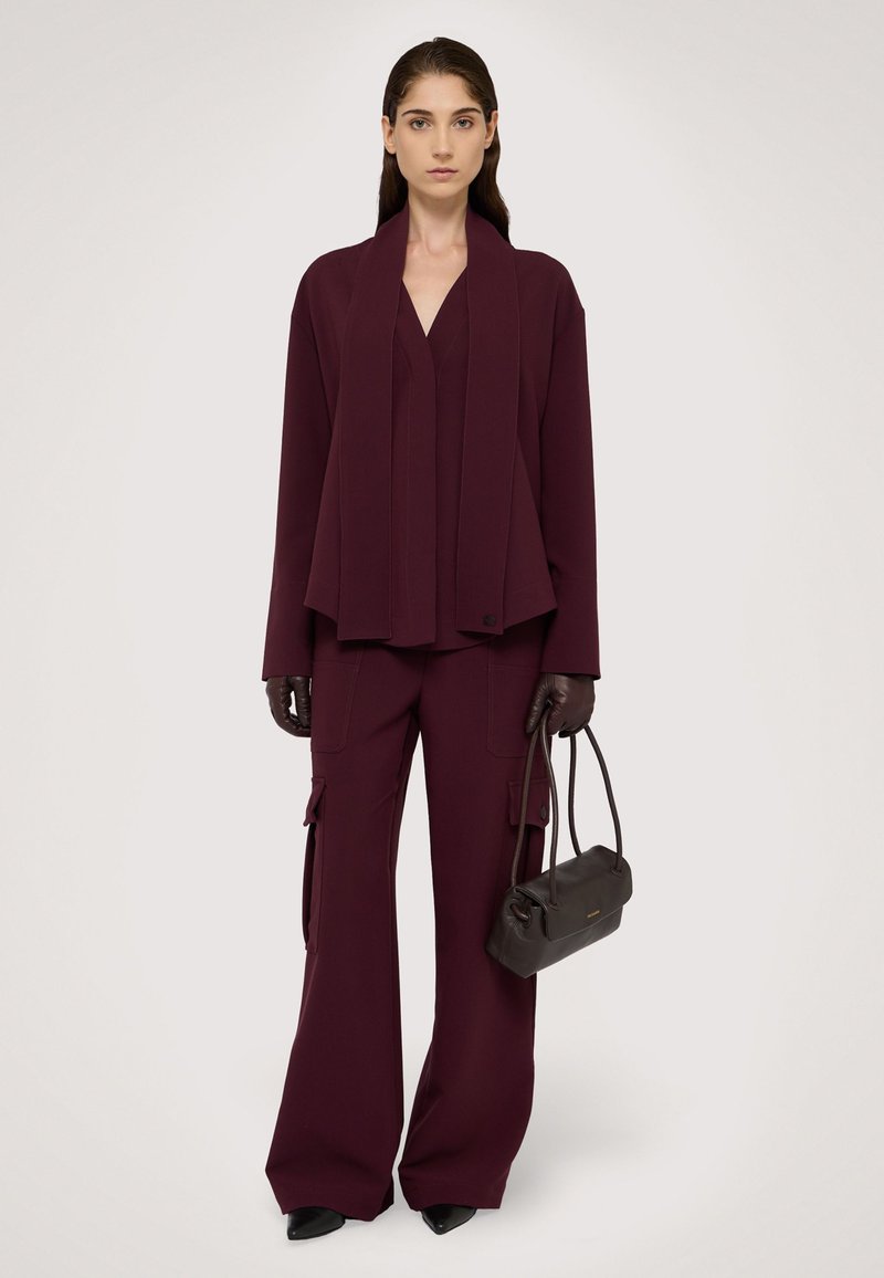 Burgundowy garnitur skrojony na miarę z szerokimi spodniami, drapowana marynarka i top z głębokim dekoltem w serek. Dopełnia go mała ciemna torebka i rękawiczki.
