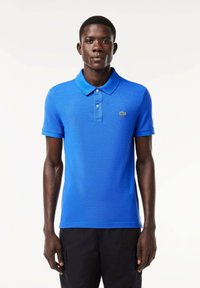 Camisa polo azul de algodón con cuello clásico, dos botones, mangas cortas y un pequeño logo verde de Lacoste en el lado izquierdo del pecho.