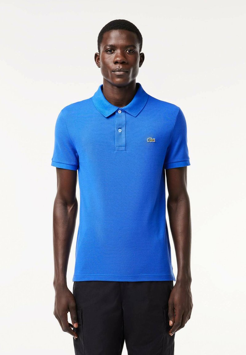Camisa polo azul de algodón con cuello clásico, dos botones, mangas cortas y un pequeño logo verde de Lacoste en el lado izquierdo del pecho.