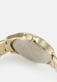 Reloj de pulsera metálico dorado con caja redonda, corona texturizada y brazalete metálico liso. Logo grabado en la corona visible.
