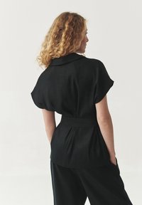Zwarte blouse met korte mouwen, een kraag, een ceintuur in de taille en een zachte textuur. Heeft een losse pasvorm en subtiele plooien aan de achterkant.