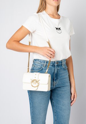 Sac à main en cuir blanc avec une fermeture à rabat, des accessoires en or et une bandoulière en chaîne. Il présente un anneau décoratif avec des accents ressemblant à des oiseaux.