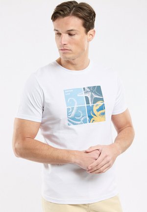 Jeune homme portant un t-shirt blanc avec un motif carré divisé en quatre sections montrant des tentacules, une étoile de mer et un crabe jaune.