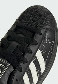 Black leather sneaker with a textured toe cap, white stripes, and a star design. Features black laces and a contrasting rubber sole.