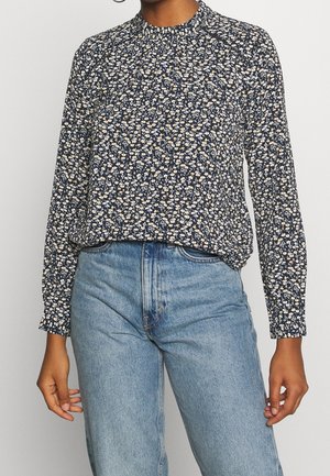 Femme portant un chemisier bleu marine à manches longues avec un petit imprimé floral blanc et un jean taille haute bleu clair.