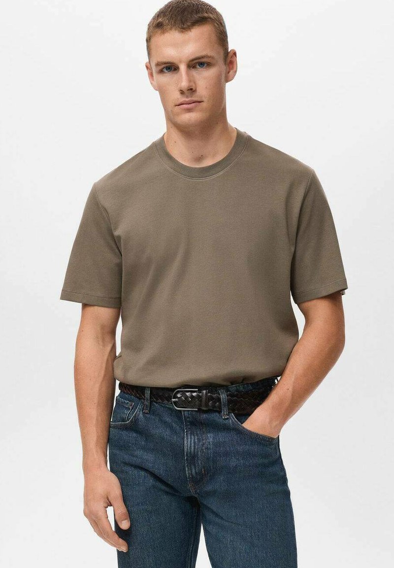 Ung mand med kort hår iført en ensfarvet olivengrøn t-shirt, der er stoppet ned i blå jeans med et sort vævet bælte, hånd i lommen.