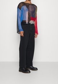 Colorful, sheer knit top with long sleeves, featuring a mix of blue, red, black, and neutral tones. Paired with black wide-leg jeans and boots.