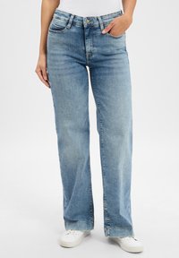 Persona vistiendo jeans rectos de color azul claro, zapatillas blancas, camiseta blanca, de pie con una mano en el bolsillo y la otra descansando al lado.