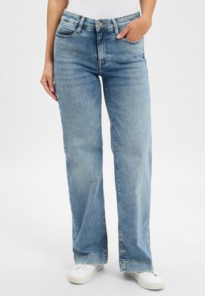 Person trägt hellblaue gerade Jeans, weiße Sneakers und ein weißes Oberteil. Er steht mit einer Hand in der Tasche und die andere Hand entspannt an der Seite.
