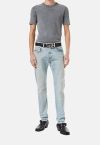 Gray ribbed short-sleeve shirt, light blue denim jeans, black leather belt with silver buckle, black clogs, standing against a white background.