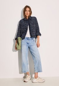 Giacca camicia blu navy a trama con due tasche, t-shirt bianca, jeans a gamba larga azzurri chiari e sneakers beige. Tieni un oggetto lavorato a maglia verde.