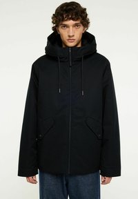 Ung man med lockigt hår, iförd en svart huva-jacka med framfickor och dragkedja, ihop med blå jeans, mot en neutral bakgrund.