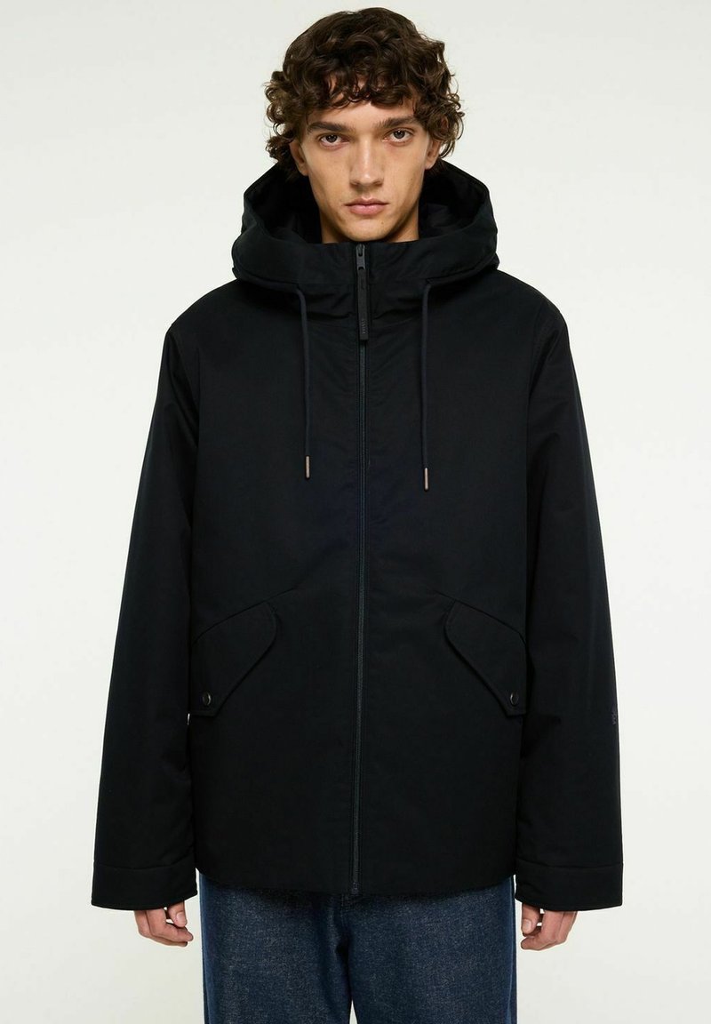 Ung man med lockigt hår, iförd en svart huva-jacka med framfickor och dragkedja, ihop med blå jeans, mot en neutral bakgrund.