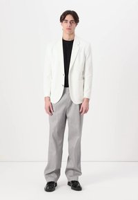 White blazer with a single-breasted design, two front pockets, worn over a black shirt. Light gray dress pants and black shoes complete the outfit.