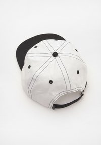 Fehér sapka fekete karimával és állítható pánttal. Fekete pöttyökkel és kontrasztos varrással rendelkezik. Pamutból készült, strukturált dizájnnal.