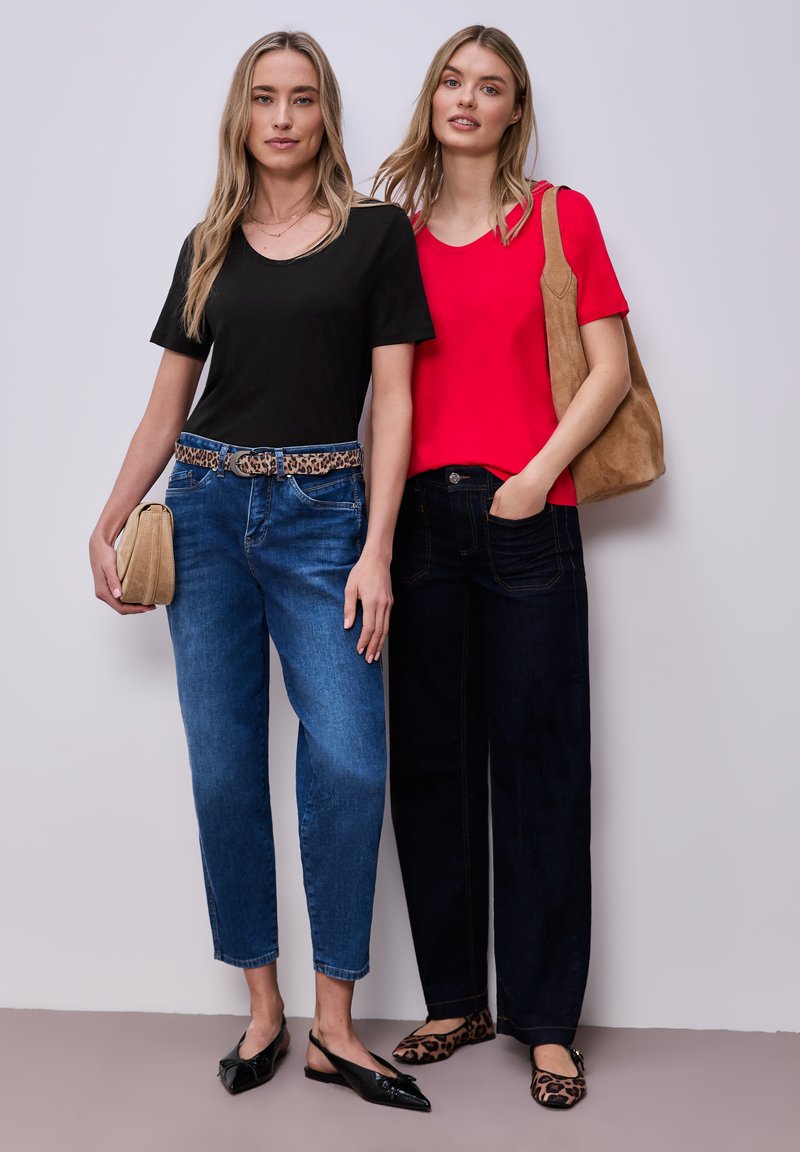 Deux femmes se tiennent côte à côte, portant des hauts décontractés, des jeans et des accessoires à motif léopard, l'une tenant une pochette beige et l'autre un sac beige.