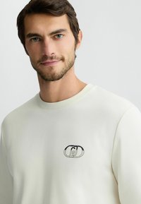 Crème-kleurige sweatshirt met ronde halslijn, ribgebreide manchetten en een klein zwart logo geborduurd op de linkerkant van de borst. Glad textuur.