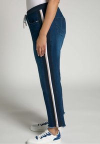 Pantalones jogger de mezclilla en azul oscuro, con franjas laterales en negro y blanco. Cintura elástica con cordón; detalle dedo en el bajo desgastado.