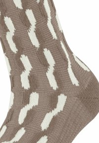 Nahaufnahme einer gestrickten Socke mit einem beigen und weißen, sich wiederholenden abstrakten Muster sowie einer strukturierten Ferse und Sohle.