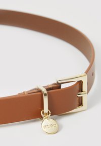 Cintura in pelle marrone con fibbia quadrata dorata e un ciondolo rotondo inciso con "HUGO." Texture liscia, design minimalista.