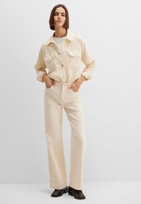 Beige Cord-Hemdjacke mit zwei Brusttaschen, kombiniert mit passenden hoch taillierten Hosen. Das Modell trägt schwarze Schuhe; glatter Hintergrund.