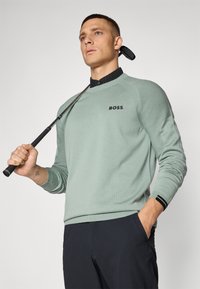 Lysegrønn genser med teksturerte ermer og svart logo "BOSS," brukt sammen med en svart skjorte med krage. Modellen holder en golfklubbe.