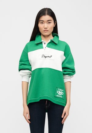 Femme aux longs cheveux noirs portant un sweat-shirt à col vert et blanc avec des logos, debout devant un fond gris clair uni.
