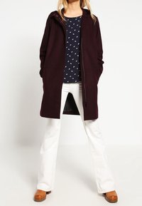 Cappotto bordeaux con cappuccio, chiusura lampo intera e tasche laterali, indossato sopra una maglietta blu navy con motivi bianchi. Abbinato a pantaloni bianchi a zampa d'elefante.
