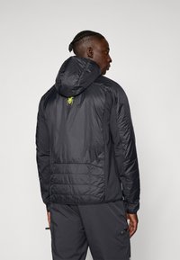 Melna, viegla, siltināta jaka ar kapuci. Iezīmēta ar izšūtu muguru un dzeltenu zirneklis logotipu, kas atrodas centrā augšējā muguras daļā.