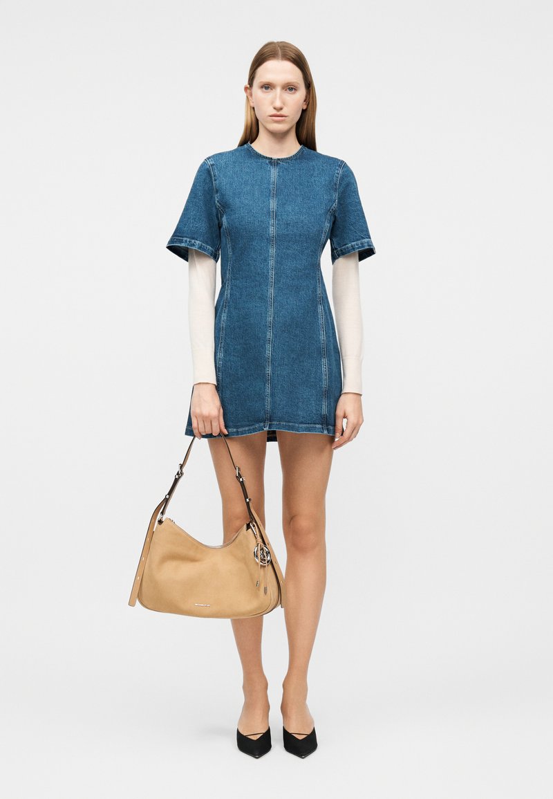 Γυναίκα με κοντομάνικο φόρεμα από τζιν, φορεμένο πάνω από μακρυμάνικη λευκή μπλούζα, κρατώντας μια μπεζ τσάντα και φορώντας μαύρα μυτερά παπούτσια.