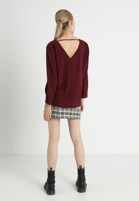 Top bordeaux a maniche lunghe con scollo a V sul retro abbinato a una minigonna tartan e stivali neri alla caviglia, realizzato con un tessuto testurizzato.