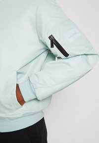 Ljus mintgrön bomberjacka med en zippered ärmlomma, ribbade muddar och en slät, glänsande yta. Varumärkeslogotyp på ärmen.