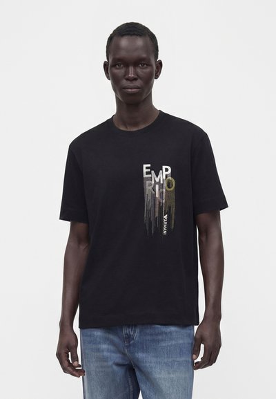 Άνδρας που φοράει μαύρο T-shirt Emporio Armani με κάθετο σχεδιασμό κειμένου και μπλε τζιν, στέκεται μπροστά σε ένα απλό λευκό φόντο.