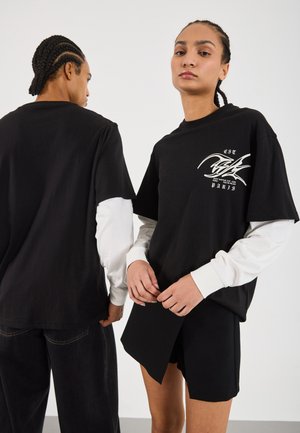 Black oversized t-shirt with white graphic design, layered over a long-sleeve white shirt. Black shorts complete the outfit, with textured fabric.