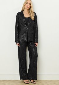 Donna con lunghi capelli biondi, che indossa un blazer di paillettes nere e pantaloni a gamba larga, in piedi contro un muro bianco semplice e un pavimento in legno.
