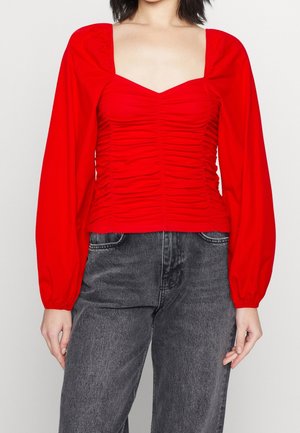 Nainen, joka käyttää punaista röyhelöityä pitkähihaista toppia, jossa on neliskanttinen pääntie, ja mustia korkeavyötäröisiä farkkuja, seisoo yksiväristä taustaa vasten.