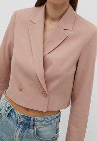 Cropped blazer in soft pink fabric, featuring a notched collar, double-button closure, and smooth texture. Paired with high-waisted jeans.