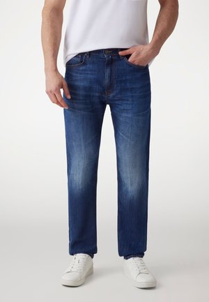 Men's blue denim jeans with a straight-leg design, faded wash, five pockets, and a zipper fly, paired with white sneakers.