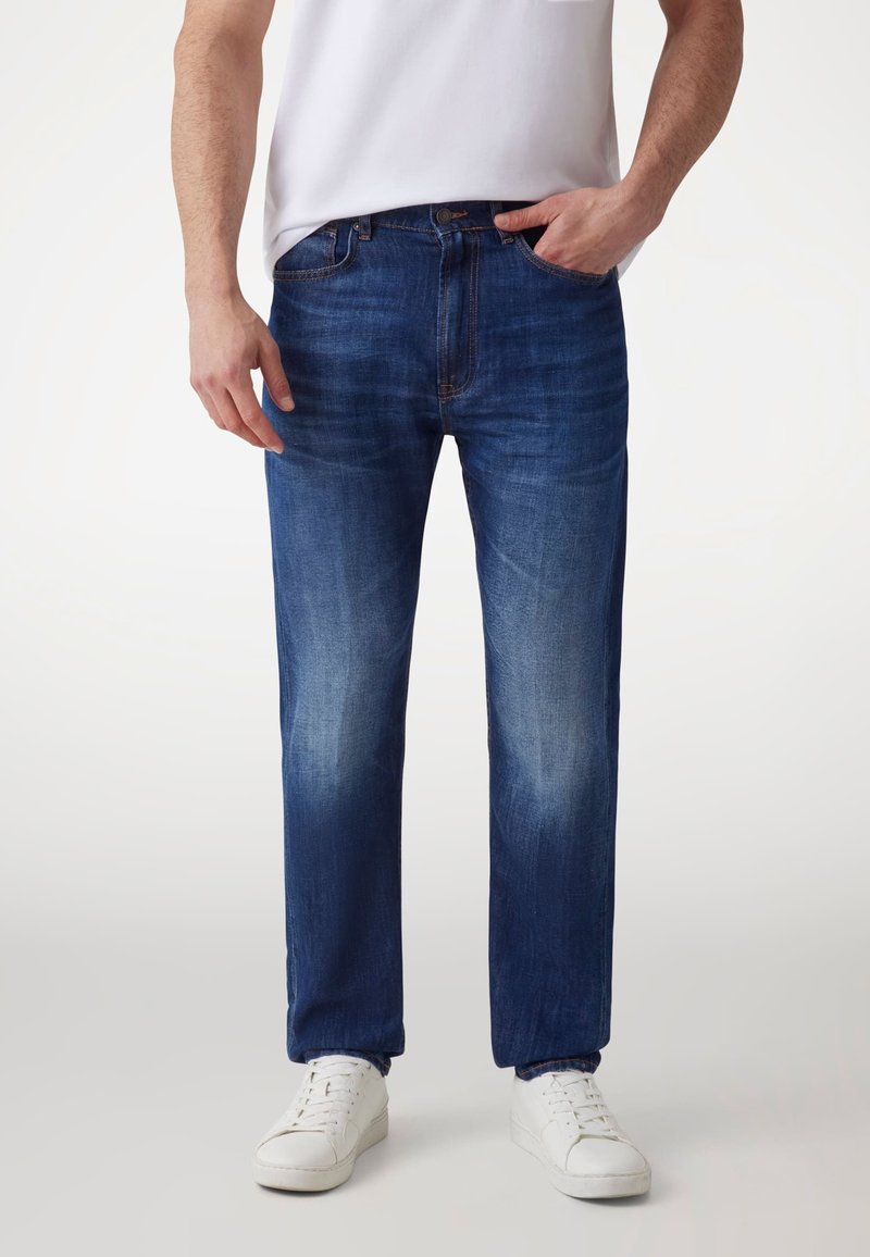 Jeans de mezclilla azul para hombre con un diseño de pierna recta, lavado desgastado, cinco bolsillos y cierre con cremallera, combinados con zapatillas blancas.