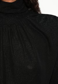 Crni vrh visokog ovratnika s glitterom, sakupljenim detaljima tkanine blizu ovratnika, koji nosi osoba s kovrčavom tamnom kosom.