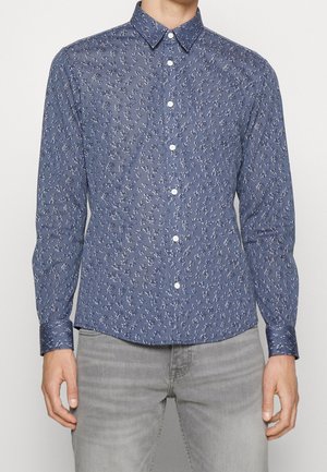 Mann trägt ein eng anliegendes blaues Hemd mit kleinem weißen Blumenmuster und hellgraue Jeans, steht vor einem schlichten Hintergrund.