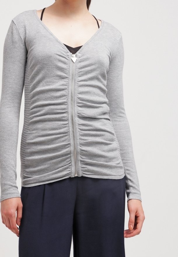Cardigan zippé gris avec un col en V, doté d'une texture froncée sur le devant et de manches longues. Porté par-dessus un haut noir.