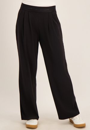 Zwarte wijde broek met plooien in de taille gedragen met witte sneakers op een lichte achtergrond.